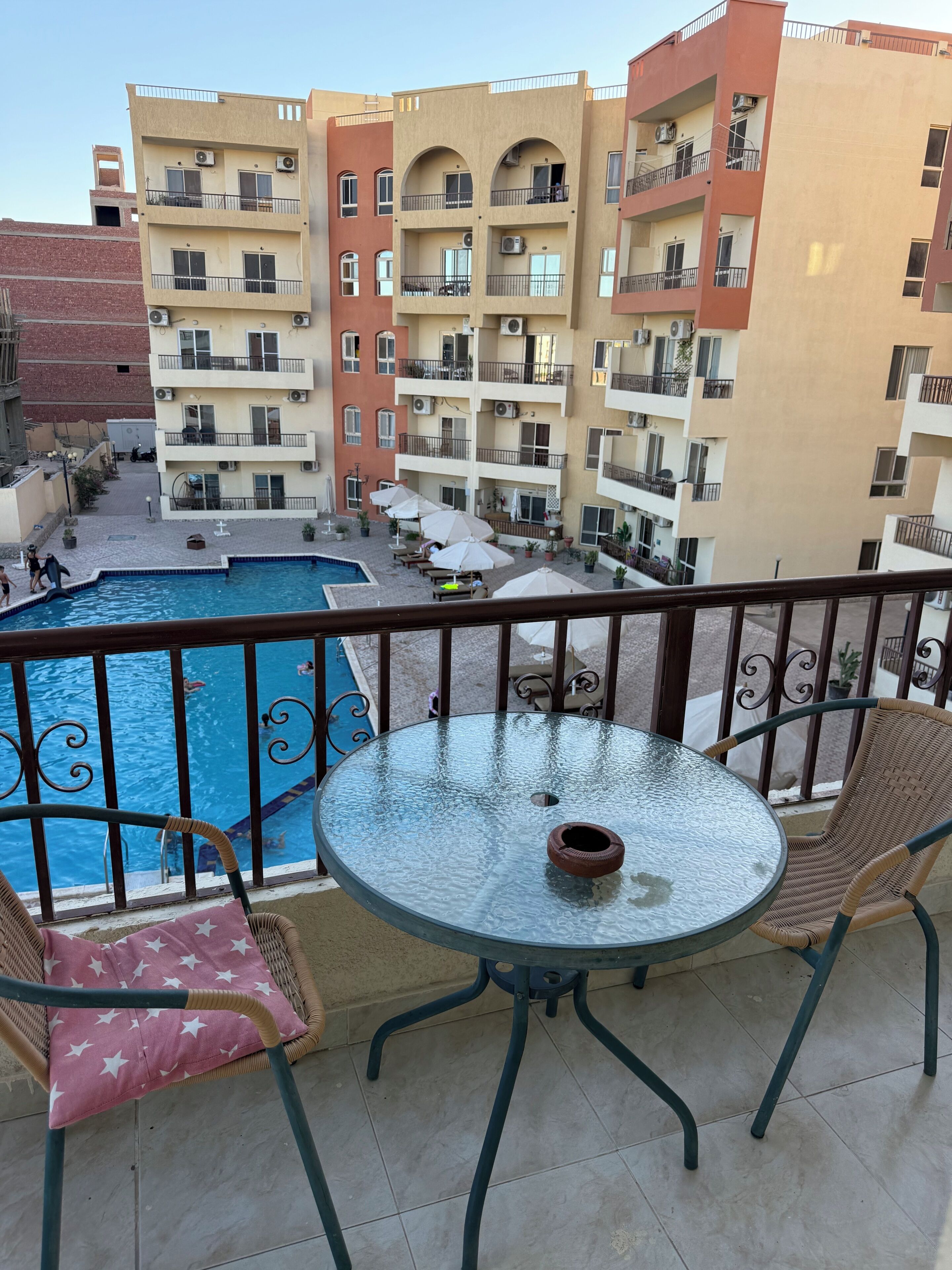 A balcony with a glass table, wicker chairs, and a view of a swimming pool and multi-story buildings.