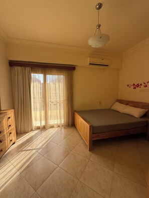 A bedroom with a bed, a wooden dresser, a window with sheer curtains, and an air conditioning unit.