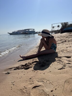 A person sitting on a sandy beach near the water, with a boat in the background. - Apartment in the center of Hurghada 7 min walk from the beach (Hurghada)