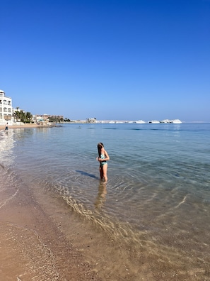 A beach scene with clear shallow water, a person in a swimsuit, and boats in the distance.