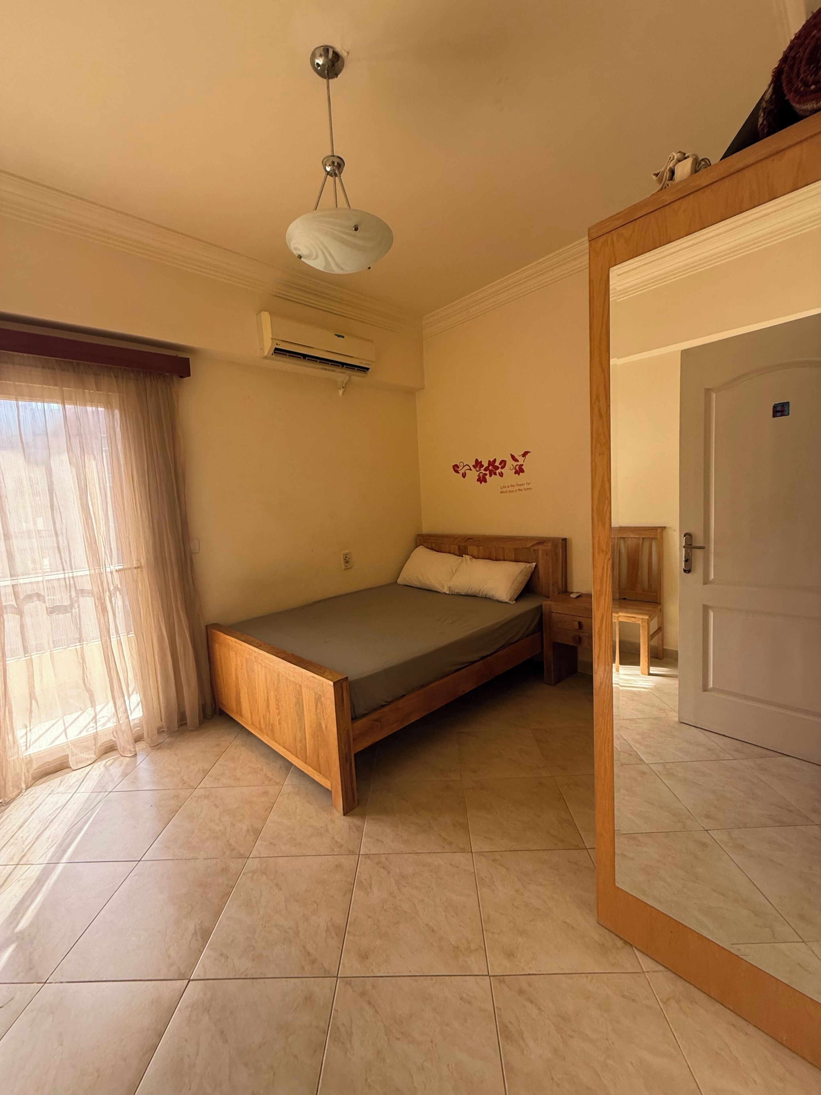 A bedroom with a bed, a wooden headboard, a ceiling light, an air conditioning unit, and a door.