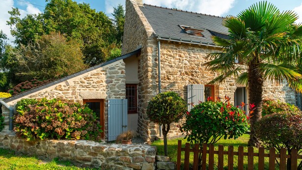 Una casa de piedra con techo inclinado, cerca de madera y un pequeño jardín con diversas plantas y una palmera.