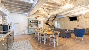A spacious living area with a wooden dining table, white chairs, a blue armchair, and a staircase leading to an upper level. - Gite La Petite Ferme (Clermont-les-Fermes)