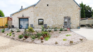 A stone building with a sloped roof, a large door, and a small window. There are outdoor chairs and a garden with various plants and flowers. - Gite La Petite Ferme (Clermont-les-Fermes)