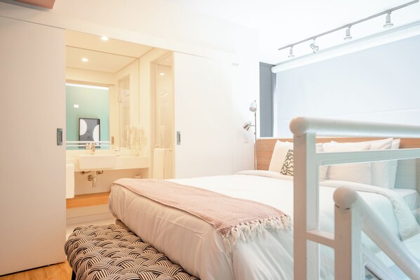 Un dormitorio moderno con cama, banco y baño visible a través de una puerta abierta.