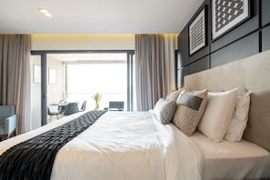 Quarto de hotel moderno com uma cama grande, um sofá e uma área de refeições visível através de uma porta aberta.