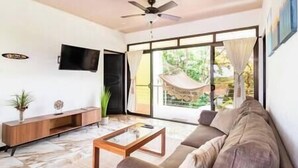 A living room with a sofa, coffee table, TV, and a large window with a view of greenery. - Deluxe Penhouse with Pool - 3 min walk to the beach (Playa Pelada)