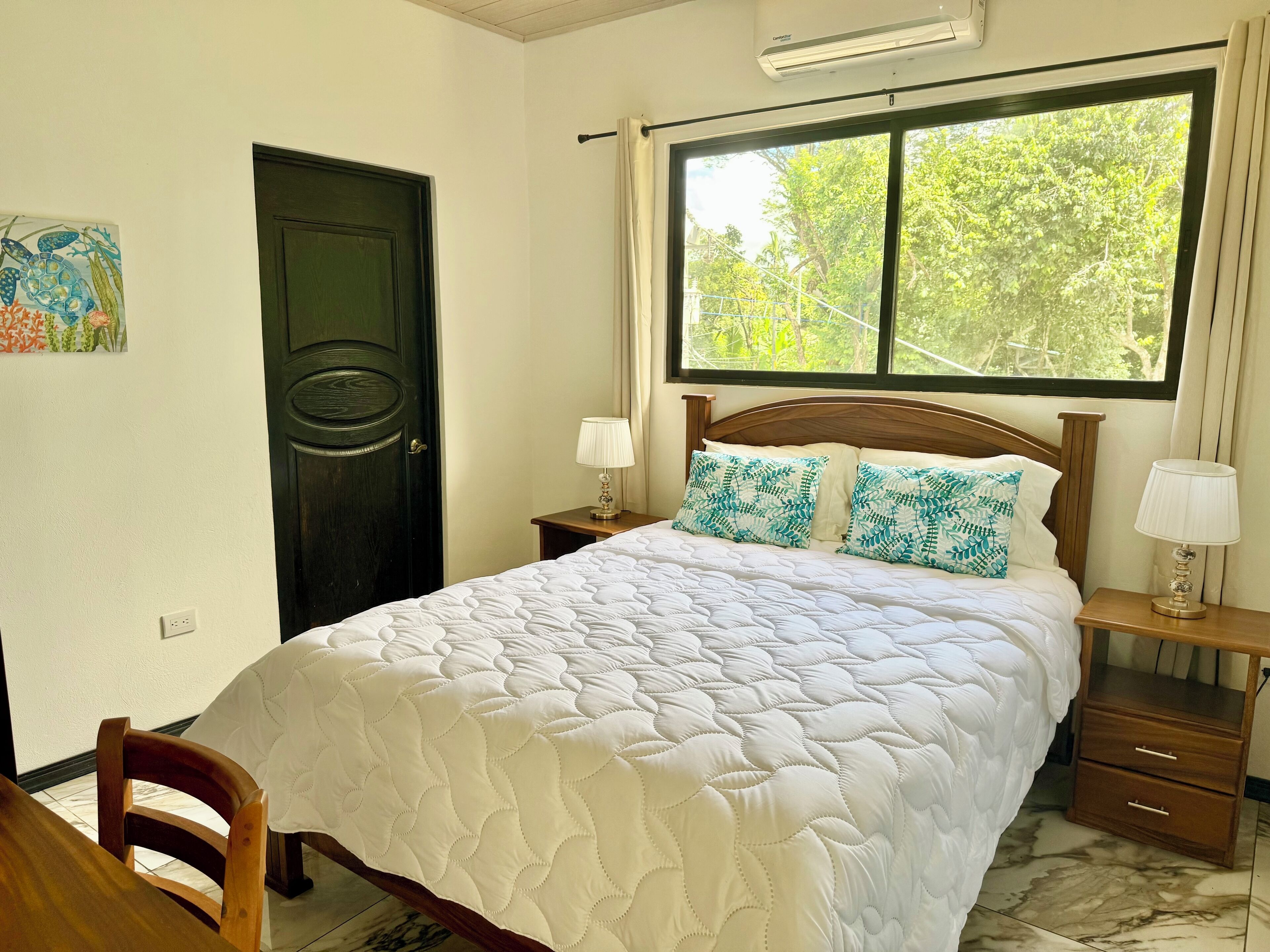 A bedroom with a bed, two bedside tables, a nightstand, a chair, a door, and a window with a view of trees.