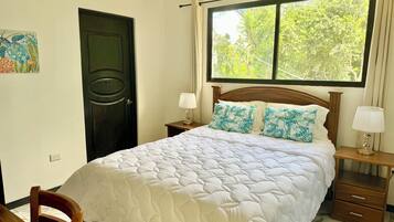 A bedroom with a bed, two bedside tables, a nightstand, a chair, a door, and a window with a view of trees.
