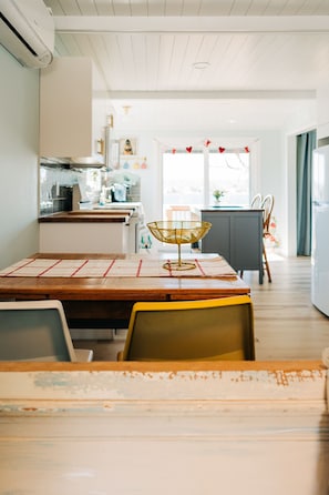 Un comedor con mesa de madera, sillas y una cesta decorativa. Se ve una cocina al fondo, con una ventana y vistas al exterior.