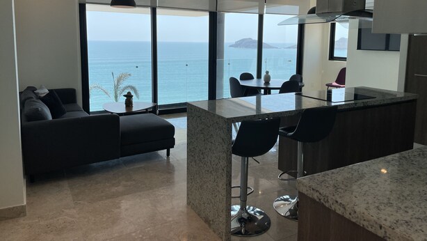 A modern kitchen with a large island, bar stools, and a view of the sea. - Central 102 Apartments (Mazatlán)