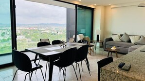 A modern living room with a large window offering a city view, a dining area with a table and chairs, and a sofa with cushions. - Central 102 Apartments (Mazatlán)