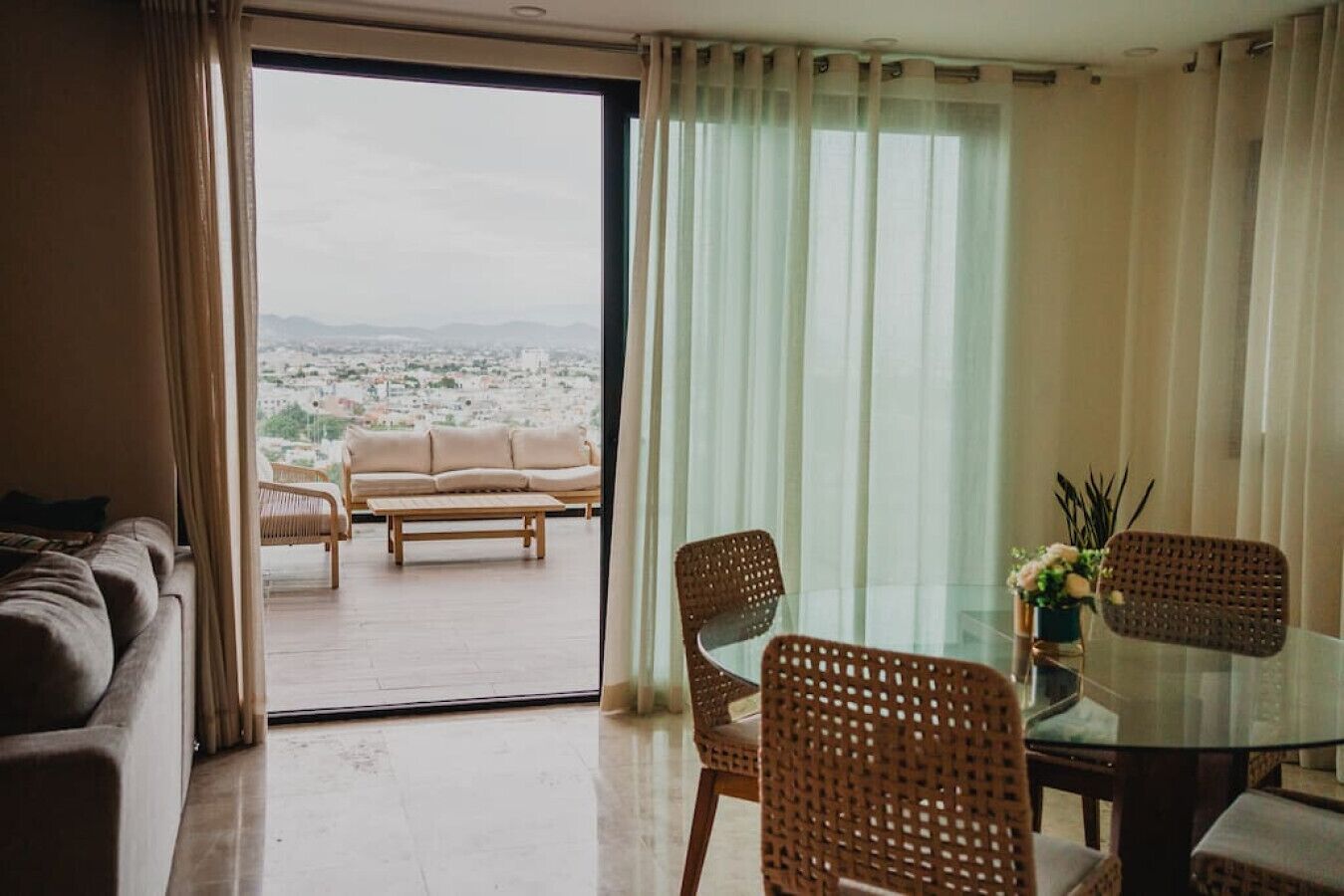 Una sala de estar moderna con una mesa de comedor de vidrio, sillas de mimbre, un sofá y vistas a un paisaje urbano a través de un ventanal amplio.