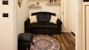 Una habitación con un sofá negro, un tapete redondo y suelo de madera. Se observa un armario blanco con estantes sobre el sofá.