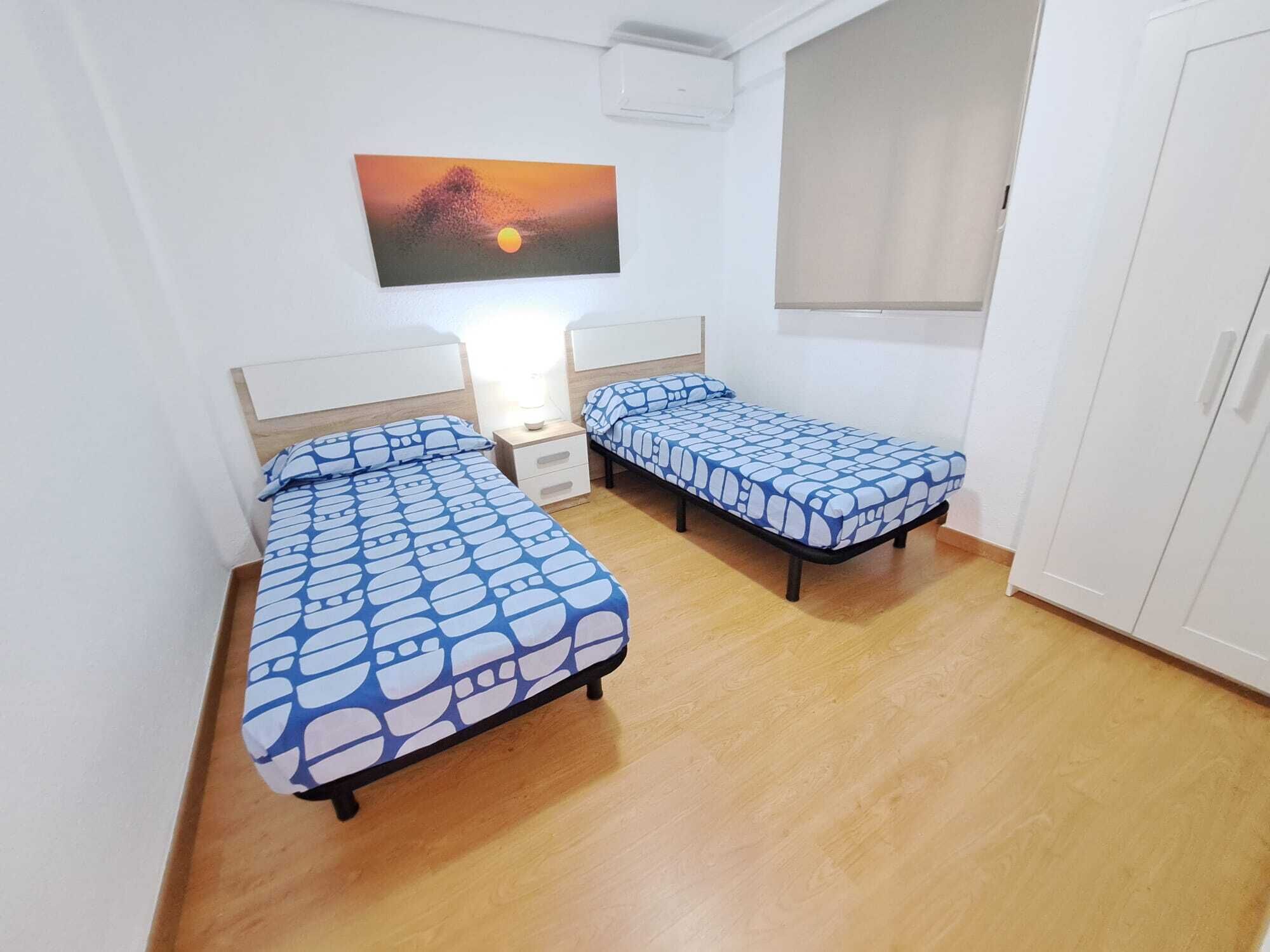 A small room with two single beds, a nightstand, a wall-mounted air conditioner, and a window with a blind.