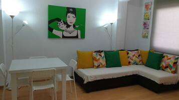 Un área de estar con un sofá blanco, cojines de colores, una mesa blanca y sillas. En la pared, un cuadro verde.
