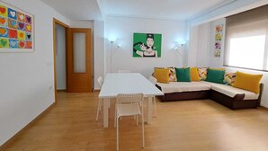 Een moderne woonkamer met een witte eettafel, een bank met kleurrijke kussens, en een schilderij aan de muur.