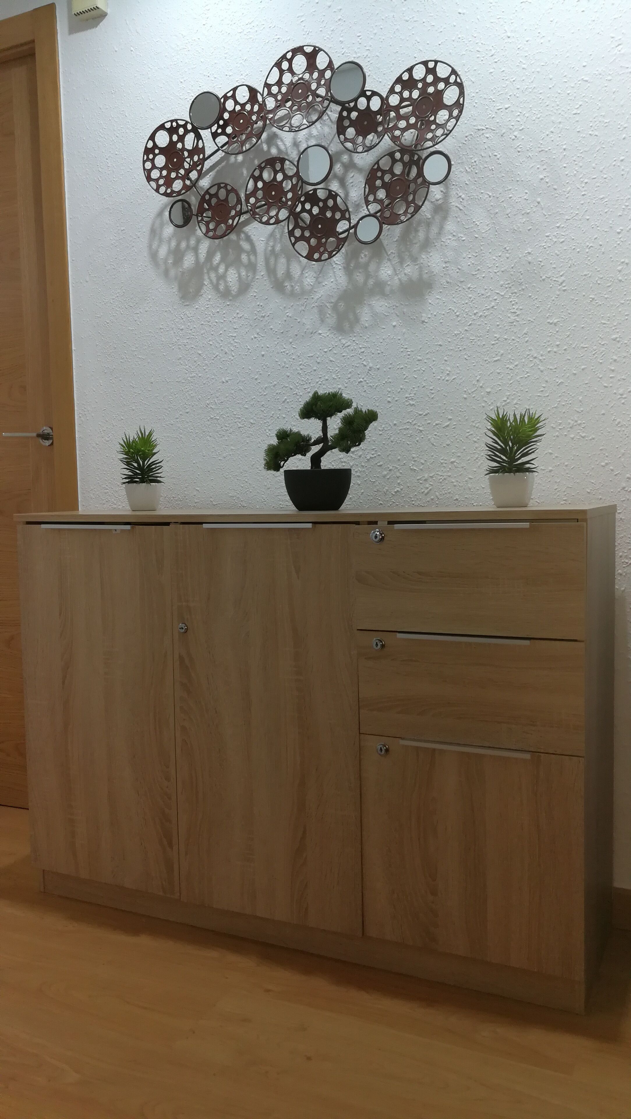 Ett träskåp med två krukväxter ovanpå, en dekorativ väggdekoration och en dörr till vänster.
