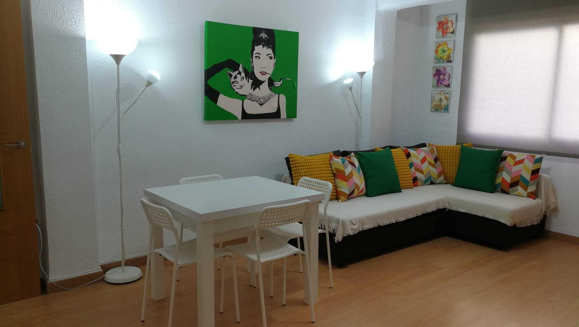 Een woonkamer met een witte bank, kleurrijke kussens, een witte eettafel met stoelen en een schilderij aan de muur.