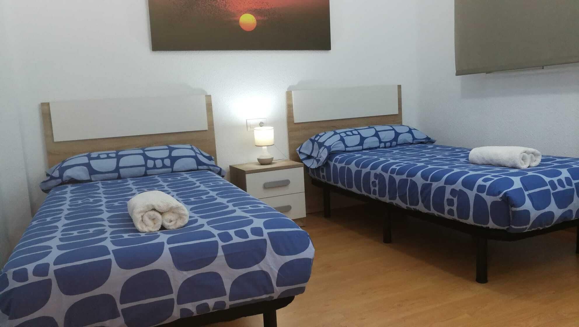 Habitación de hotel con dos camas, cada una con una colcha y almohadas a juego de diseño azul. Entre las camas, un mesita de noche con una lámpara.