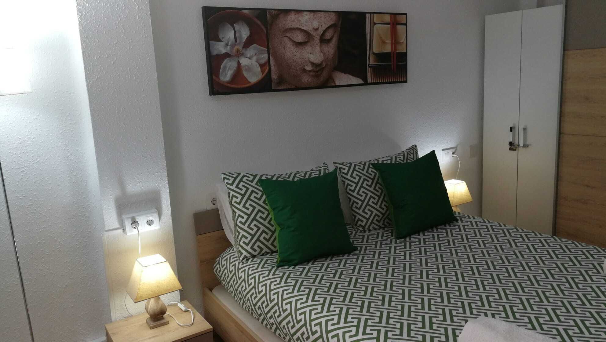 A neatly arranged bedroom with a patterned bedspread, two green pillows, a wall-mounted artwork, a bedside lamp, and a wardrobe.
