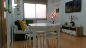 Een moderne woonkamer met een witte eettafel, witte stoelen, een bank met kleurrijke kussens, een dressoir en een groot raam met jaloezieën.