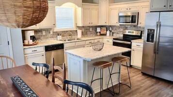 A modern kitchen with white cabinets, a marble countertop, stainless steel appliances, and a wooden dining table with bar stools.
