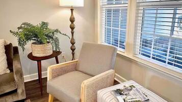A living room with a beige armchair, a small round table with a lamp, a potted plant, and a window with blinds.