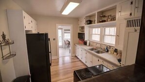 Una cocina con gabinetes blancos, un refrigerador negro, una estufa y un lavamanos. Hay una ventana sobre el lavamanos y una puerta que da a otra habitación.