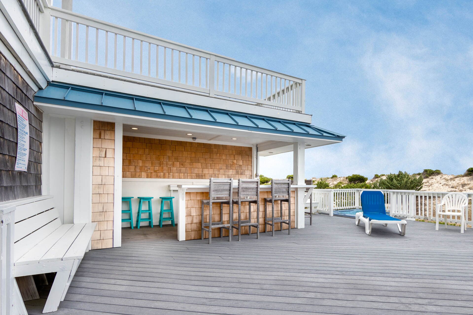Un espace extérieur abrité doté d’une terrasse en bois, d’un bar avec des tabourets et d’un coin salon au bord de la piscine avec des fauteuils.