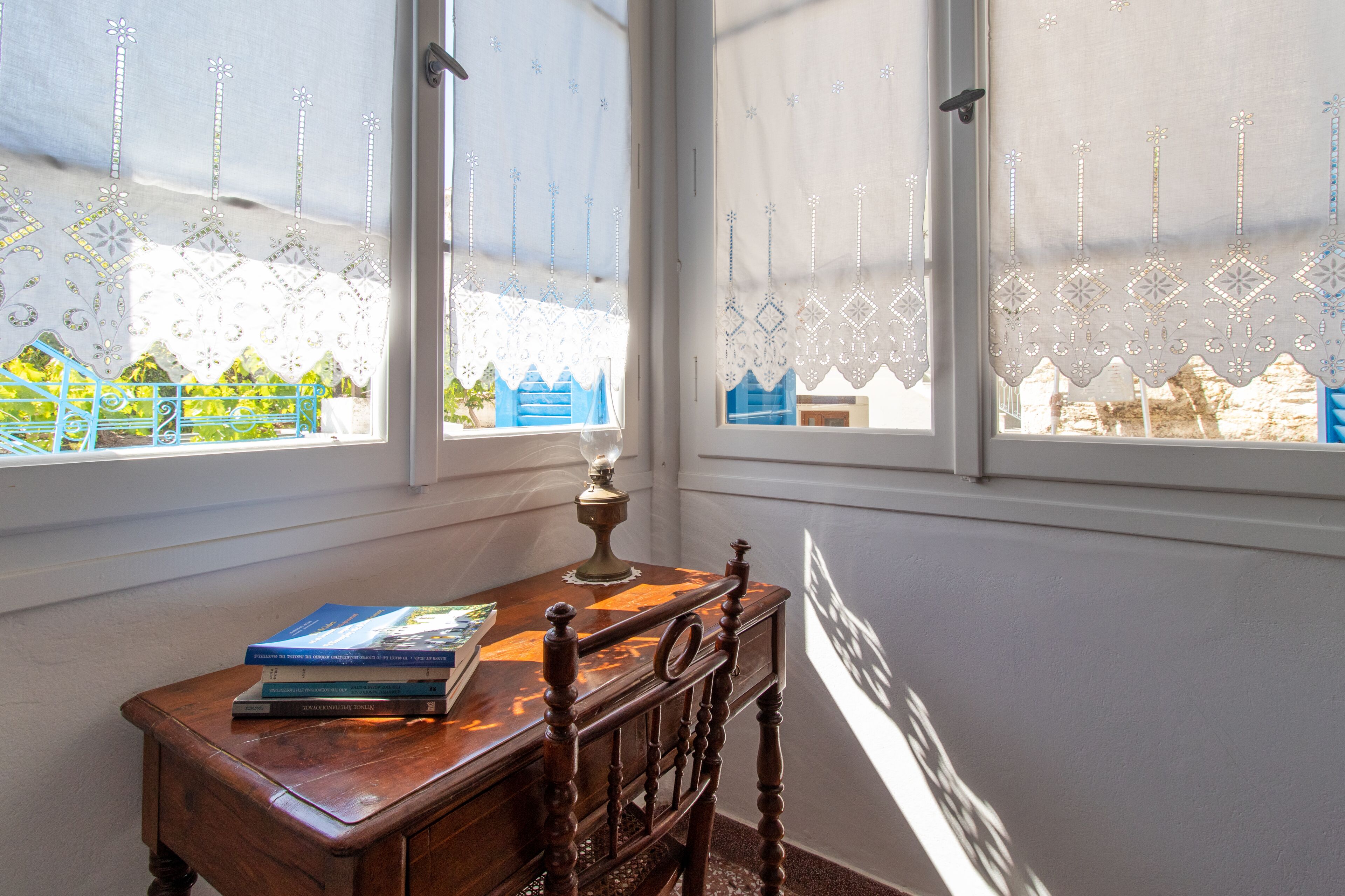 Ein Eckzimmer mit Schreibtisch und Stuhl aus Holz, einem Fenster mit Spitzenvorhängen und Blick auf ein blaues Gebäude.