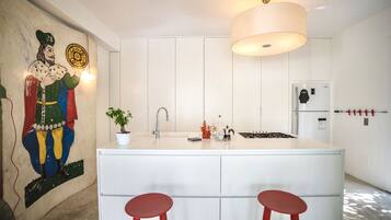 Une cuisine moderne avec des meubles blancs, un îlot central doté de tabourets, un luminaire suspendu et un réfrigérateur décoré d’un personnage de dessin animé.