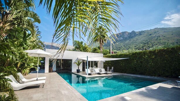 A modern outdoor pool area with lounge chairs and umbrellas, surrounded by lush greenery and a mountainous backdrop. - Modern Villa in Sicily with Pool (Cinisi)
