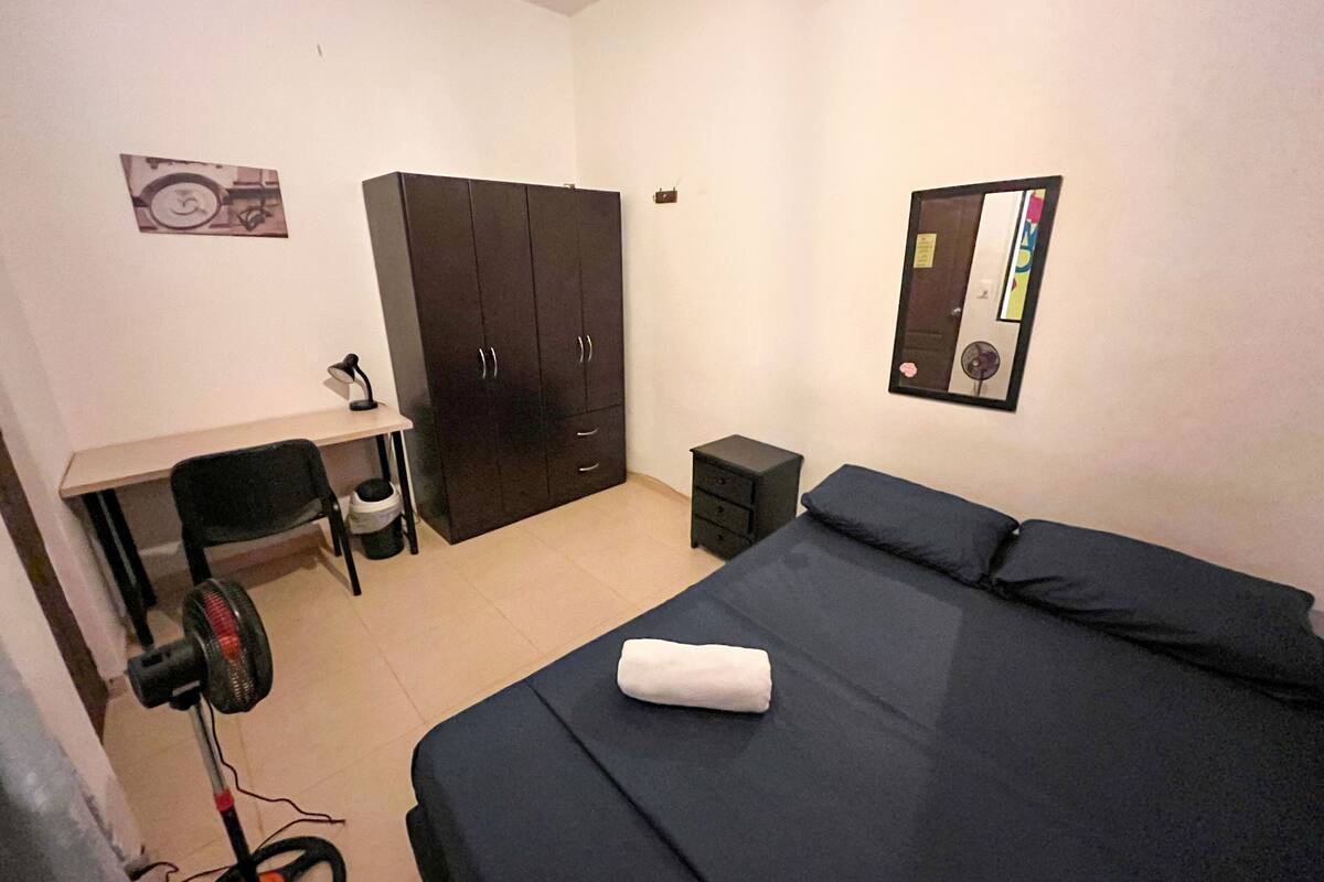 Quarto com cama, escrivaninha, cadeira, guarda-roupa, mesa de cabeceira e espelho fixado na parede.