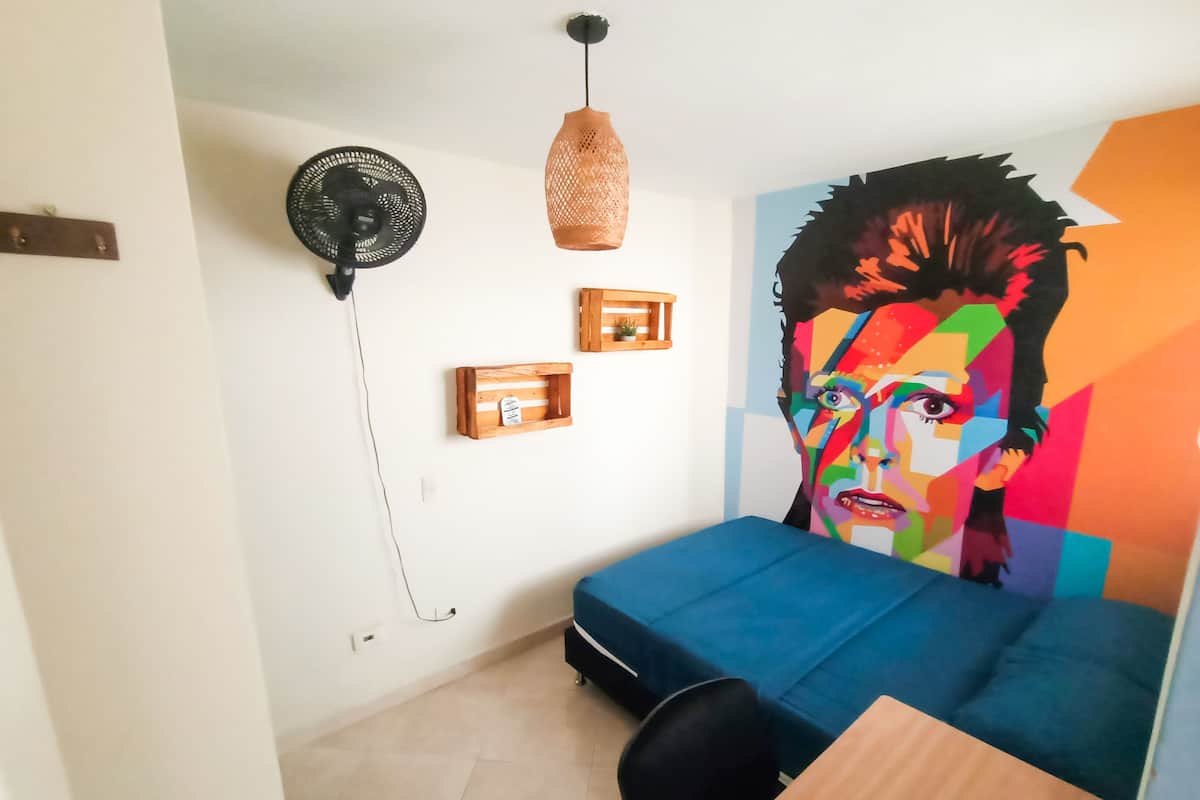 Quarto com sofá azul, mesa de madeira, cadeira preta, ventoinha na parede e pintura abstrata colorida.