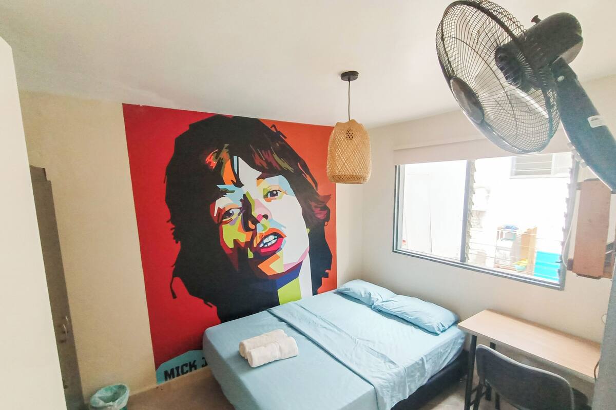 Quarto com cama, escrivaninha, janela e parede com um mural colorido.