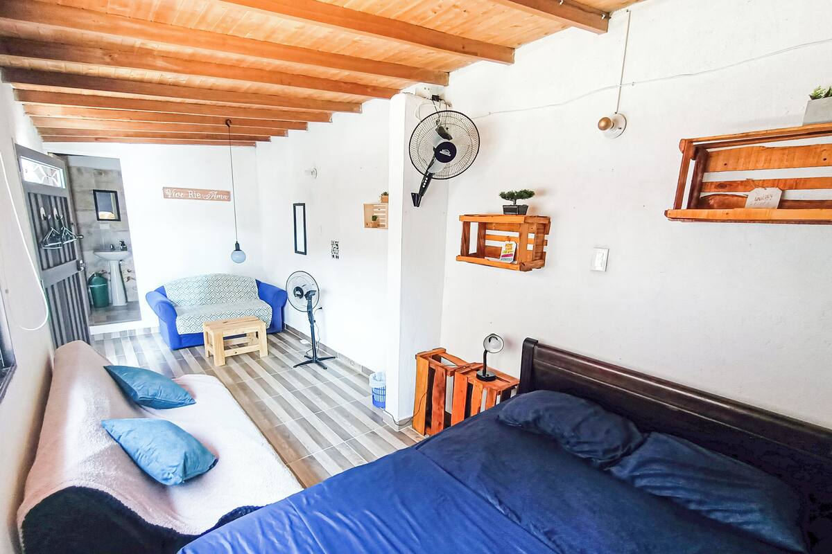 Quarto com uma cama grande, um sofá menor, um banco de madeira e uma escrivaninha com uma cadeira. Há duas ventoinhas, uma pequena prateleira com uma planta e uma placa na parede.