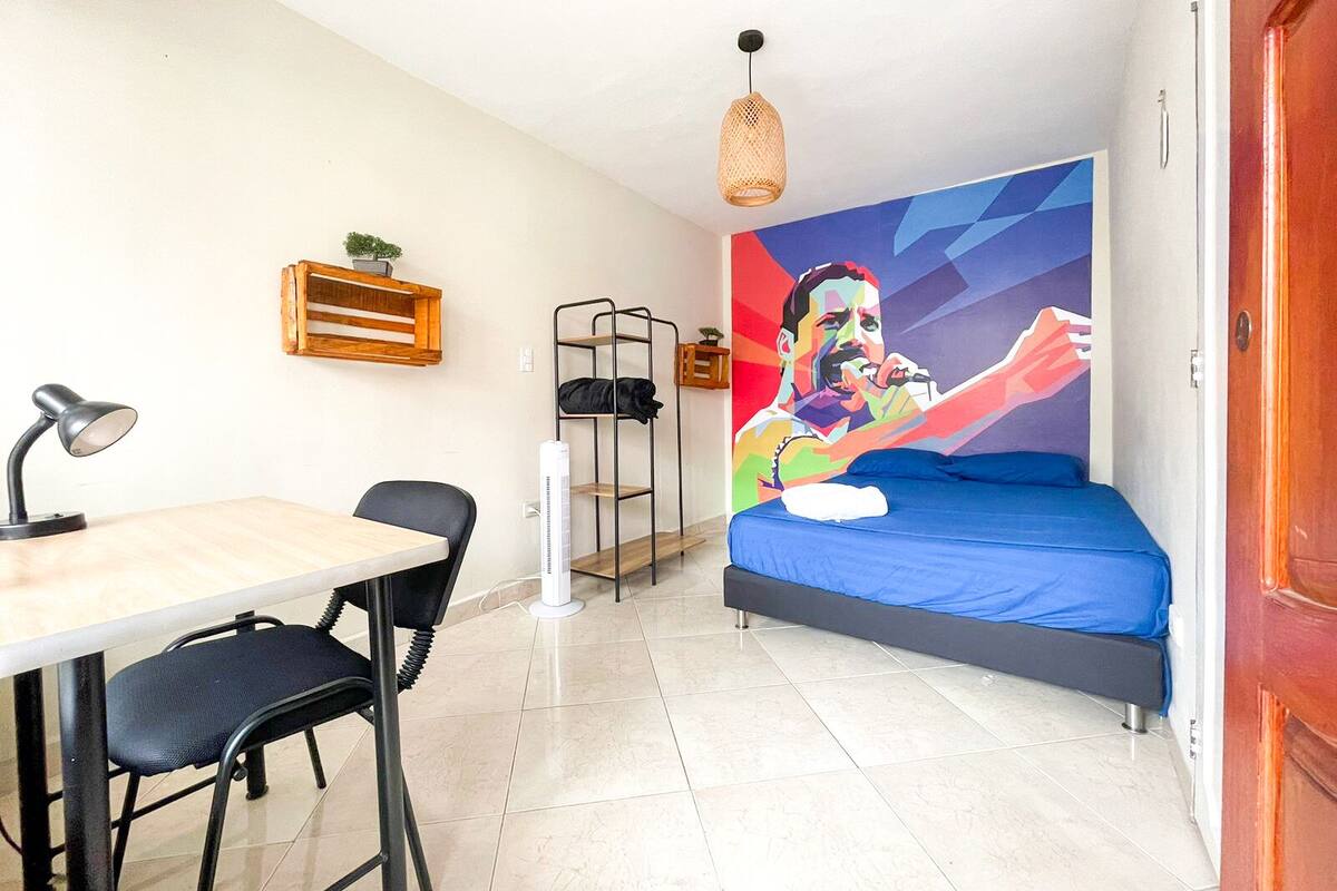 Quarto compacto com cama, escrivaninha com cadeira, aquecedor vertical, prateleira com planta e um mural colorido na parede.