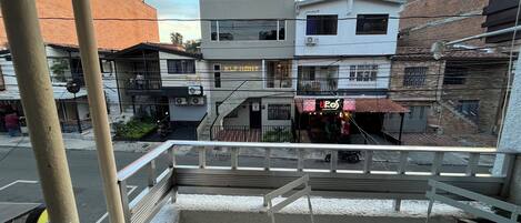 Uma varanda com mesa branca e planta em vaso, com vista para uma rua com edifícios e um restaurante chamado "UROS".