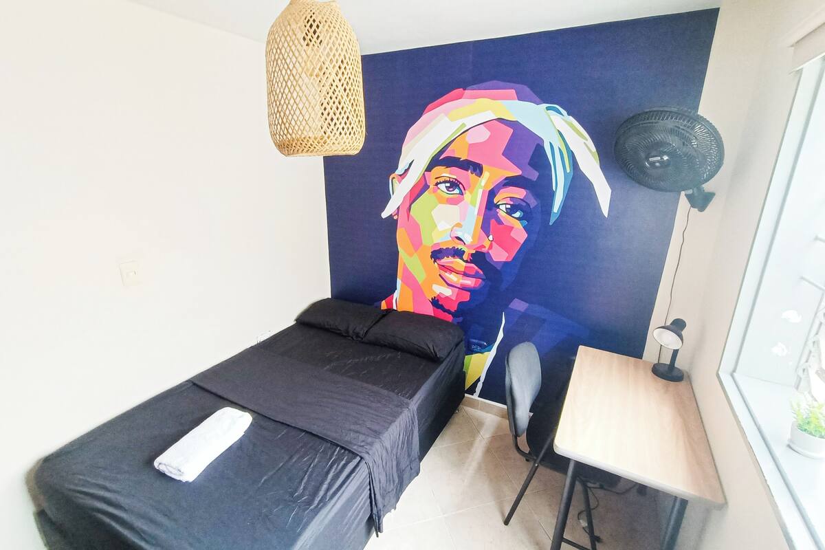 Quarto com cama, escrivaninha, cadeira, luminária pendente e ventoinha fixada na parede.
