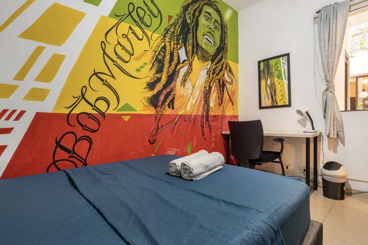 Quarto de hotel com uma cama, uma escrivaninha e uma cadeira. Há um mural colorido na parede e uma janela com cortinas.