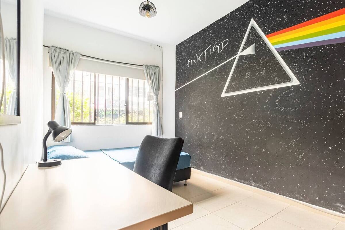 Quarto com escrivaninha, cadeira, luminária, janela com cortinas e parede com mural do Pink Floyd.