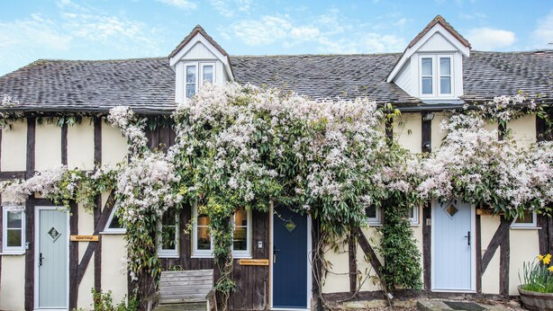 Une maison de campagne traditionnelle à colombages, avec un arbre en fleurs bien visible devant, un banc et un chemin de pierres menant à l’entrée.