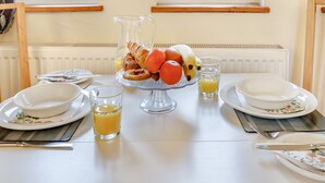 Une table de petit-déjeuner dressée avec des assiettes, un pichet de jus d’orange, un verre de jus d’orange et un saladier rempli d’oranges, de bananes et de croissants.