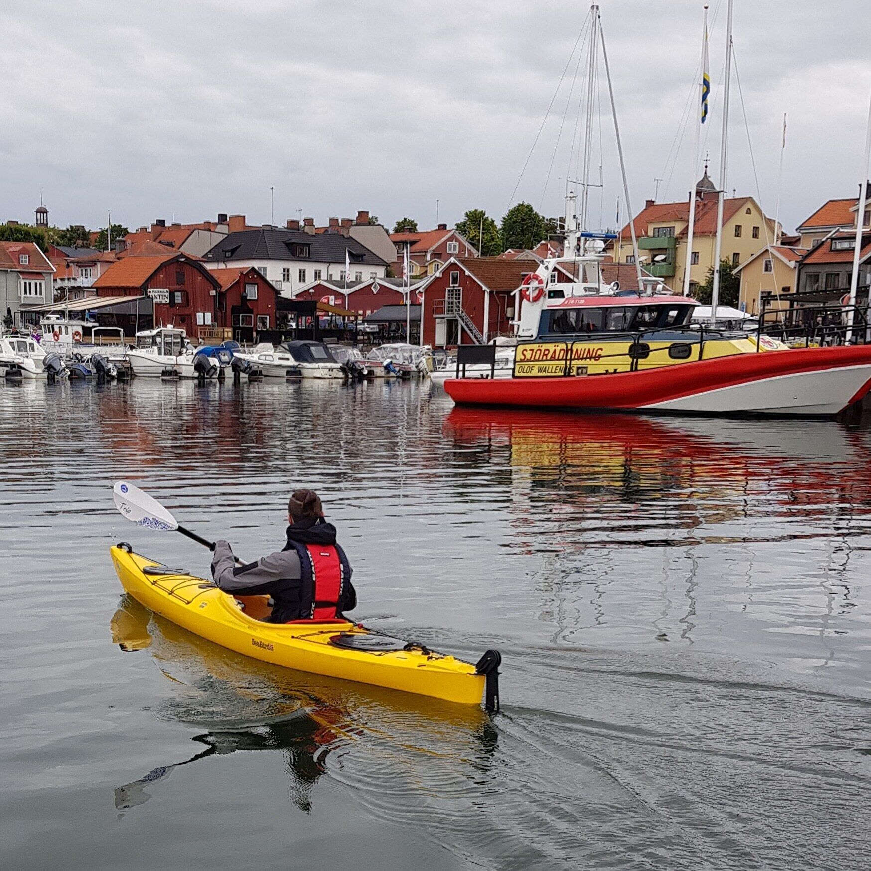 A person kayaking on calm water with a town and boats in the background.