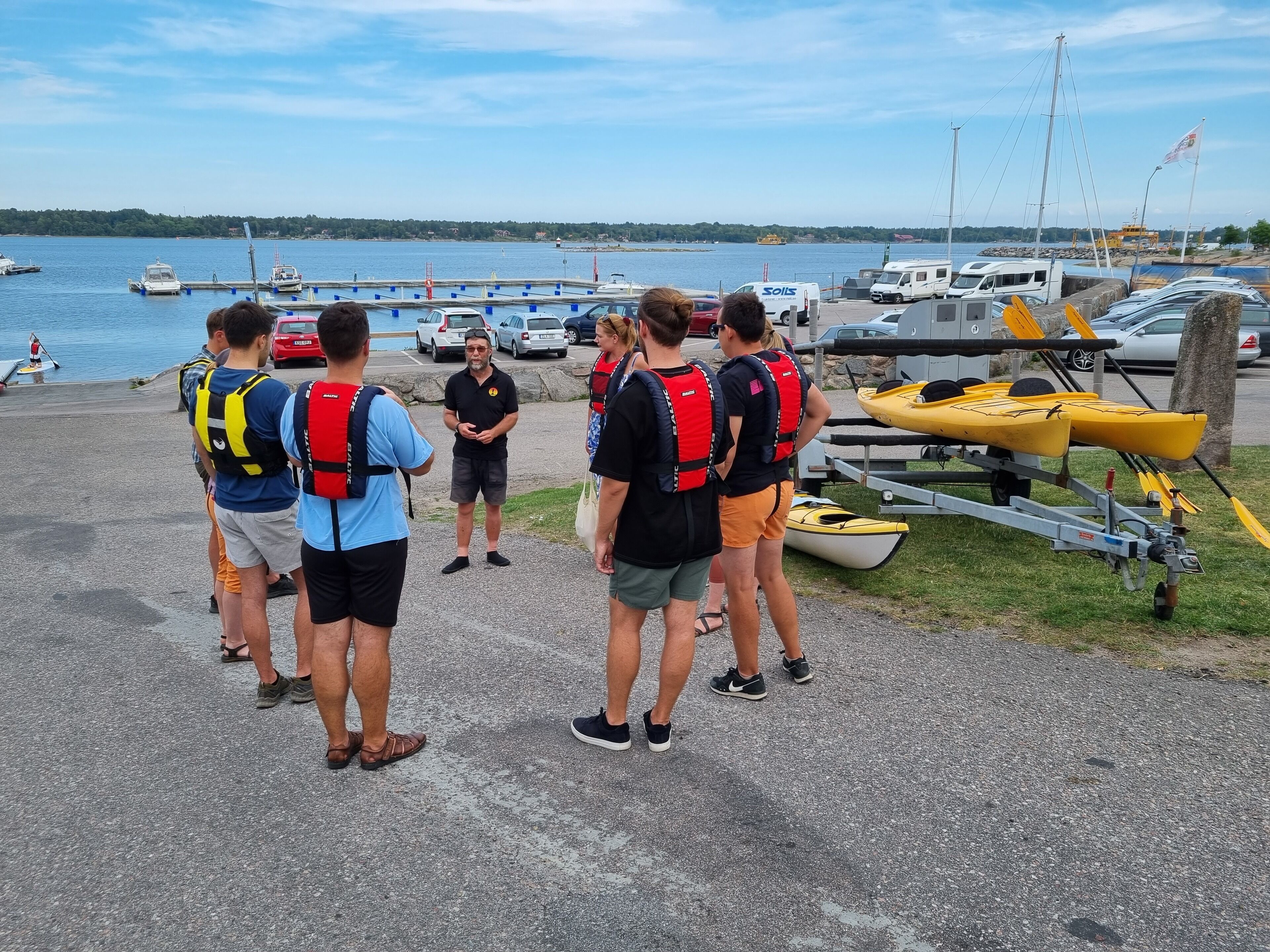 A group of people standing on a dock by a body of water, wearing life jackets, with boats and a trailer nearby.