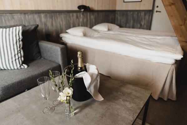 A room with a bed, a grey sofa, a table with a bottle of champagne, glasses, and a vase with flowers. - Epokgården (Öregrund)