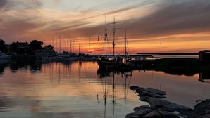 A harbor at sunset with several sailboats docked, calm water reflecting the sky, and rocky shoreline. - Epokgården (Öregrund)