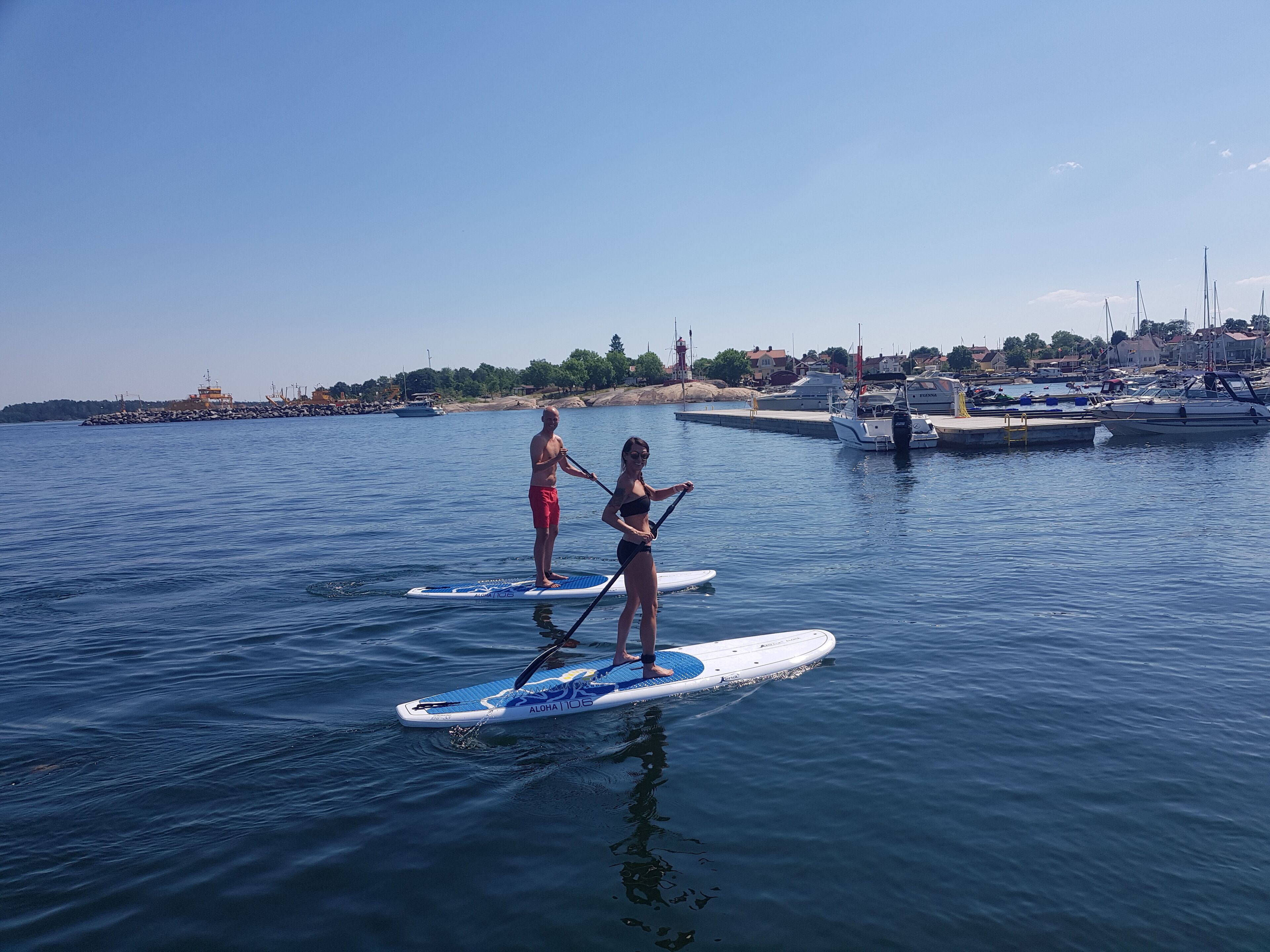 Two people paddleboarding on a calm body of water with a marina and boats in the background.