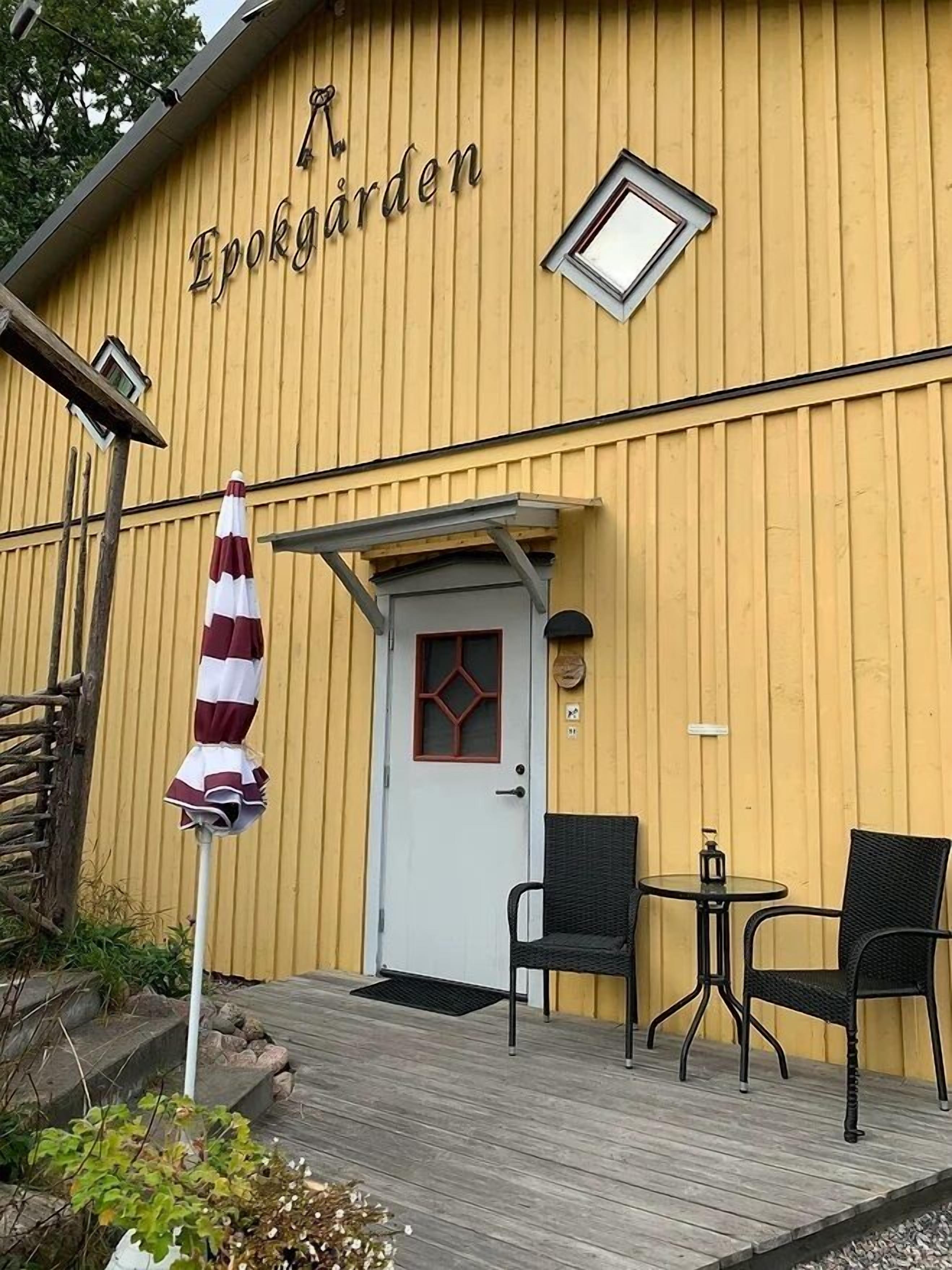 Un bâtiment jaune avec l’inscription « Epokgården » sur le mur, une porte blanche et une petite table avec deux chaises à l’extérieur.